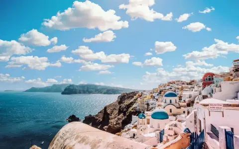 Santorini, Grecja. Białe domy z niebieskimi kopułami kościołów na zboczu góry, nad turkusowym morzem. Marzysz o podróży? Sprawdź, ile się leci do Grecji!