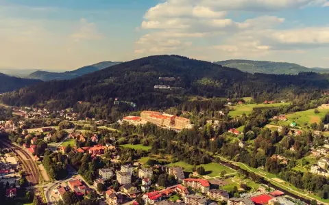 Widok z kamery na hotel Gołębiewski w Wiśle, otoczony zielonymi wzgórzami i zabudowaniami.