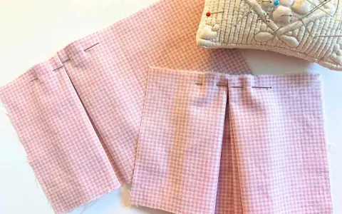 Tissu rose à carreaux avec des plis épinglés, montrant comment faire des plis en couture avec une fourchette. Coussin à épingles en arrière-plan.