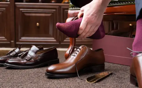 Hombre se pone calcetín morado para ablandar zapatos con crema hidratante. Zapatos de vestir y mocasines en el suelo.