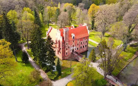 Zamek na wodzie w Wojnowicach otoczony zielenią i drzewami. Czerwona bryła zamku z charakterystycznym dachem wznosi się nad fosą.