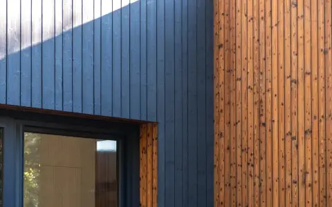 Drewniana elewacja z ciemnogranatową wiatroizolacją i naturalnym drewnem. Okno z ciemną ramą.