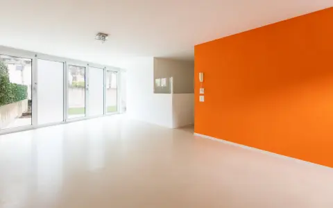 Jasne, nowoczesne wnętrze z pomarańczową ścianą i podłogą z **mikrobetonu**. Duże okna wpuszczają dużo światła.