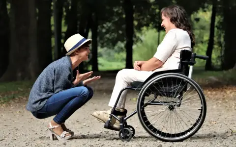 Opiekun osoby niepełnosprawnej pochyla się, by porozmawiać z kobietą na wózku inwalidzkim w parku.