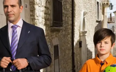 Checco Zalone con uno dei suoi figli, un bambino con un pallone da calcio, passeggiano in un vicolo.