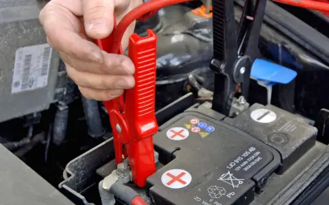 Hand klemmt rote Überbrückungsklemme an die Autobatterie, um Plus und Minus zu verbinden.