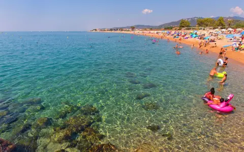 Spiaggia affollata a Santa Susanna, Spagna, con persone che nuotano e si rilassano in un mare cristallino.