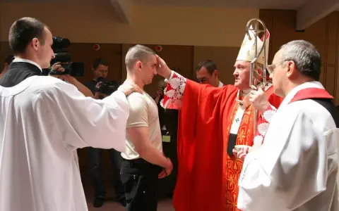 Biskup udziela sakramentu bierzmowania młodemu mężczyźnie. W tle widać osoby z kamerami.