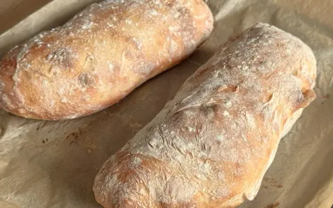 Dwa świeżo upieczone bochenki ciabatty, posypane mąką, leżą na papierze do pieczenia. Idealne do wypróbowania tego ciabatta przepis!