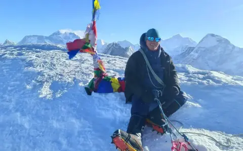 Szczęśliwy alpinista na szczycie Mera Peak, otoczony śniegiem i modlitewnymi flagami, z majestatycznymi górami w tle.
