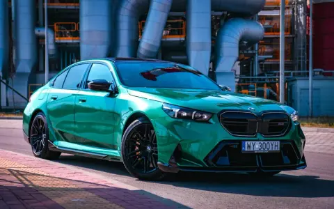Błyszczące, zielone BMW M5 G90 z czarnymi felgami stoi na tle przemysłowych rur.
