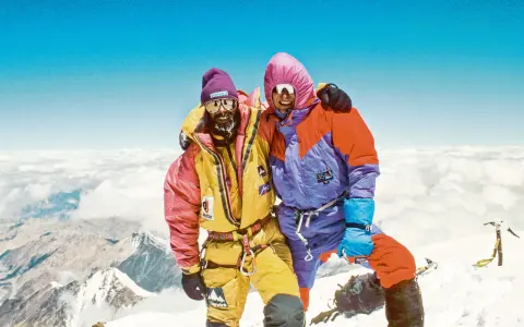 Zwei Bergsteiger auf dem Gipfel, die sich umarmen. Sie tragen bunte Daunenanzüge und Mützen. Die Frage "wie hoch ist der Mount Everest" wird hier beantwortet: 8848 Meter.