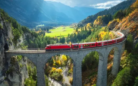 Czerwony pociąg Bernina Express przejeżdża przez kamienny wiadukt w górach jesienią.