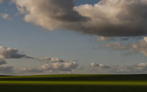 Zachód słońca nad zielonym polem, rzucającym długi cień. Chmury na niebie tworzą ciekawe fotografie.