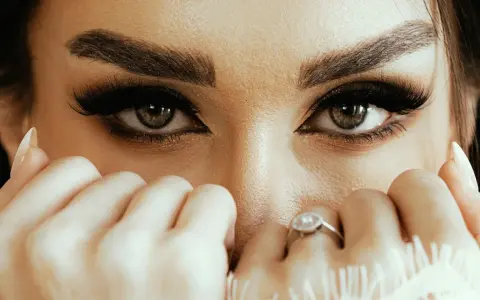 Yeux verts intenses, cils longs et maquillage sourcils parfait. Mains délicates ornées de bagues cachent une partie du visage.