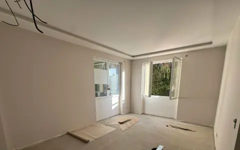Chambre en cours de rénovation avec un faux plafond élégant, deux fenêtres ouvertes sur la verdure et des matériaux de construction au sol.