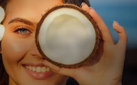 Donna sorride con un fiore tra i capelli, coprendo un occhio con una noce di cocco. Attenzione: olio di cocco sul viso fa male.