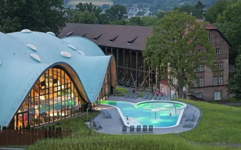 Moderne Therme mit geschwungenem Dach und beleuchtetem Pool, daneben historische Gebäude.
