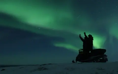 En person på snöskoter i Kiruna Lapland sträcker upp armarna mot norrskenet som lyser grönt på natthimlen.