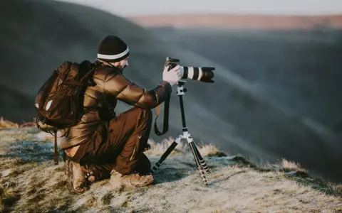 Man med ryggsäck och kamera på stativ, redo att omskola sig till naturfotograf i gryningen.