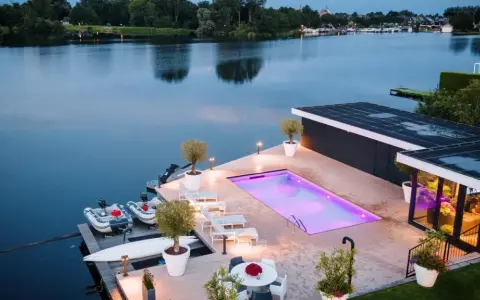 Lyxig pool med platssvetsad liner och belysning, omgiven av solstolar och växter. Båtar ligger vid bryggan.