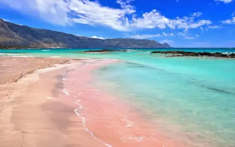 Różowy piasek plaży Elafonisi w Grecji, turkusowa woda i góry w tle.