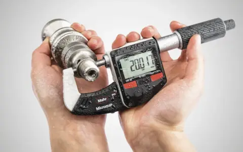 Manos sostienen un micrometro digital Mahr Micromar, midiendo una pieza metálica. La pantalla muestra 200.17 mm.