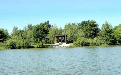 Spokojne wody magnum carp lake otaczają zielone drzewa. Mały domek z tarasem stoi przy brzegu.
