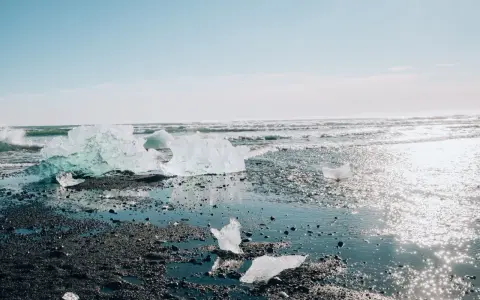 Lodowe bryły lśnią na czarnym piasku, tworząc magiczny diamond beach. Fale obmywają brzeg, a słońce odbija się w wodzie.