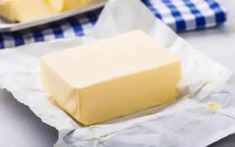Kostka masła na papierze, obok niebiesko-biała kratka. Czy masło można mrozić? Tak, można je zamrozić.