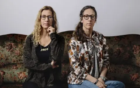 Två kvinnor sitter på en blommig soffa. Kvinnan till vänster har blont lockigt hår och ett halsband med texten "FATTA". Kvinnan till höger har mörkt hår i en hästsvans.
