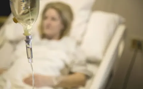 Infusion vorbereiten: Eine Frau liegt im Krankenhausbett, während eine Infusionstropfflasche und ein Tropfschlauch im Vordergrund zu sehen sind.