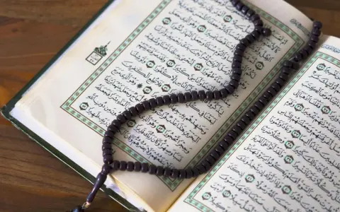 Ein Koran mit Perlenkette liegt auf einem Holztisch. Die Seiten sind mit arabischer Schrift bedruckt, die an die Geschichte von Yajuj und Majuj im Islam erinnert.
