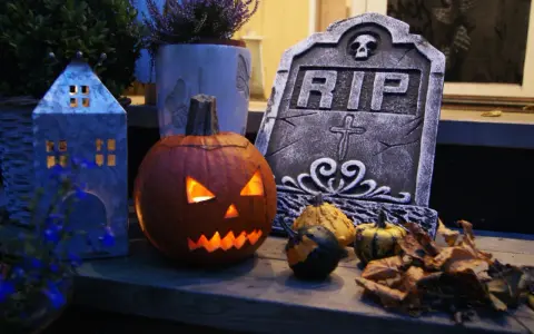 En läskig pumpa lyser upp med sitt ansikte, bredvid en gravsten och höstlöven. Perfekt för en kuslig bål halloween-kväll.