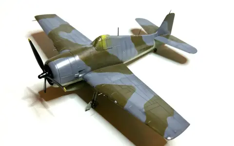Ein Modellflugzeug, ein Hellcat-Flugzeug, mit Tarnmuster in Grau und Olivgrün, steht auf weißem Hintergrund.