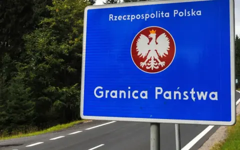 Niebieski znak z orłem i napisem "Rzeczpospolita Polska" oraz "Granica Państwa" stoi przy drodze otoczonej lasem.