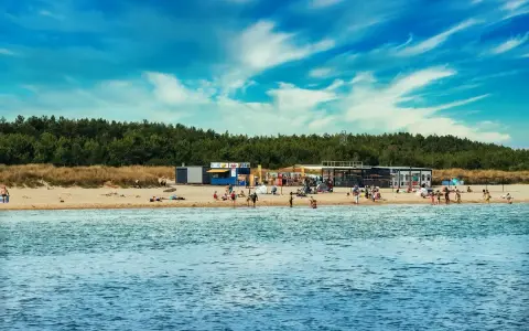 Słoneczny dzień na plaży, ludzie odpoczywają, a w tle widać budynki i las. Idealne miejsce na kemping na Helu.