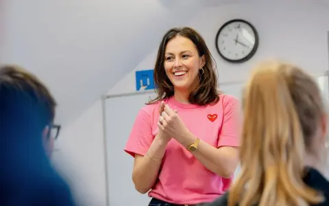 Uśmiechnięta kobieta w różowej koszulce z sercem, stojąca przed uczniami.