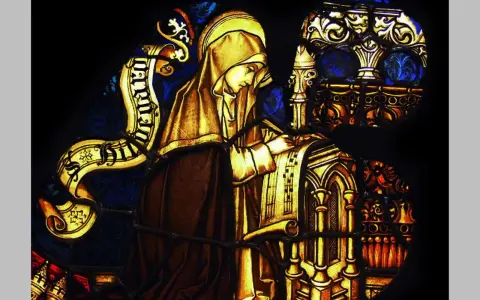 Stained glass depicts a cloaked figure, likely the head of a monastery, diligently writing on a scroll.