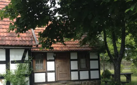 Urokliwa chata kaszubska z drewnianymi belkami i czerwoną dachówką, otoczona zielenią.