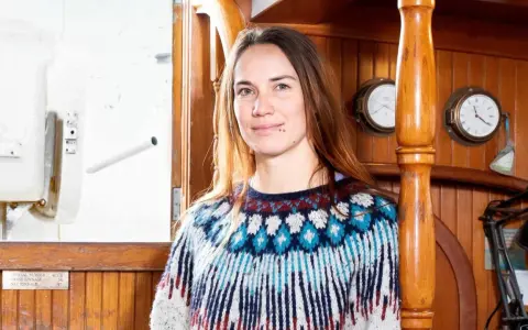 Laura Dekker heute, lächelnd in einem bunten Strickpullover, an Bord eines Schiffes mit Holzakzenten und maritimen Uhren.