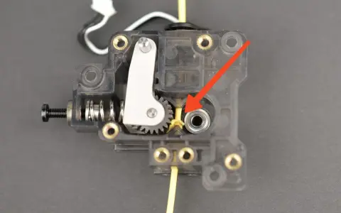 Mechanizm ekstrudera drukarki 3D, gdzie żółty filament wchodzi do mechanizmu. Strzałka wskazuje miejsce, które może wymagać czyszczenia, aby dowiedzieć się, jak wyczyścić dysze drukarki.