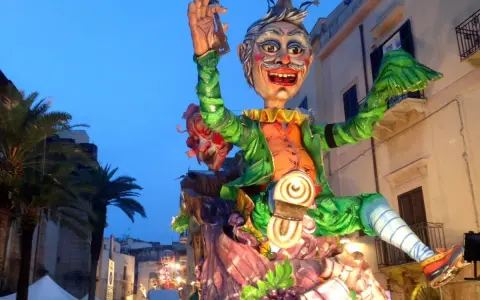 Carro allegorico con maschere di carnevale siciliane, un personaggio gioioso con vestiti verdi e arancioni sfila tra palme e palazzi illuminati.