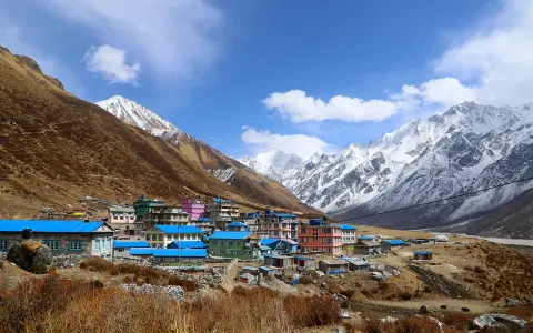 Villaggio colorato ai piedi dell'alta montagna himalayana, con case dai tetti blu e rosa, circondato da pendii bruni e cime innevate.