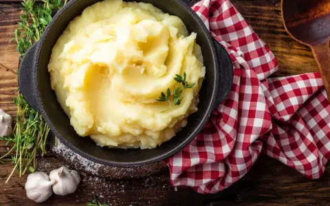 Una ciotola di cremoso purè di patate, guarnito con timo fresco, accanto ad aglio e un tovagliolo a quadri rossi.