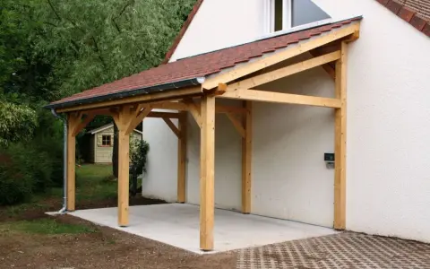 Un abri de voiture en bois avec une pente légère, adossé à une maison blanche.