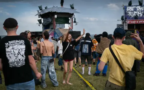 Une foule danse sous la pluie à un festival, une femme au centre lève les bras. On aperçoit une scène et des bannières, peut-être un dj français anime l'événement.