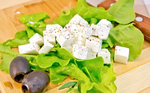 Kostki sera feta, przyprawione ziołami, leżą na liściach sałaty obok czarnych oliwek. Idealne na lekki posiłek.