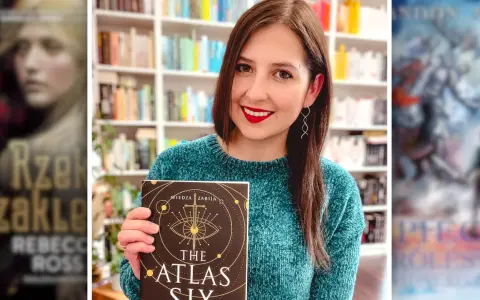 Uśmiechnięta kobieta trzyma książkę "The Atlas Six". W tle widać półki z książkami, w tym inne serie książek fantasy.