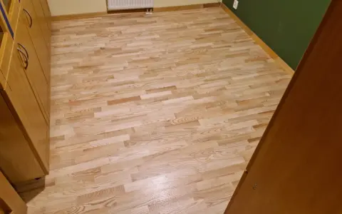 Jasny, odnowiony parkiet po cyklinowaniu, lśniący w świetle. Obok drewniane meble i zielona ściana.